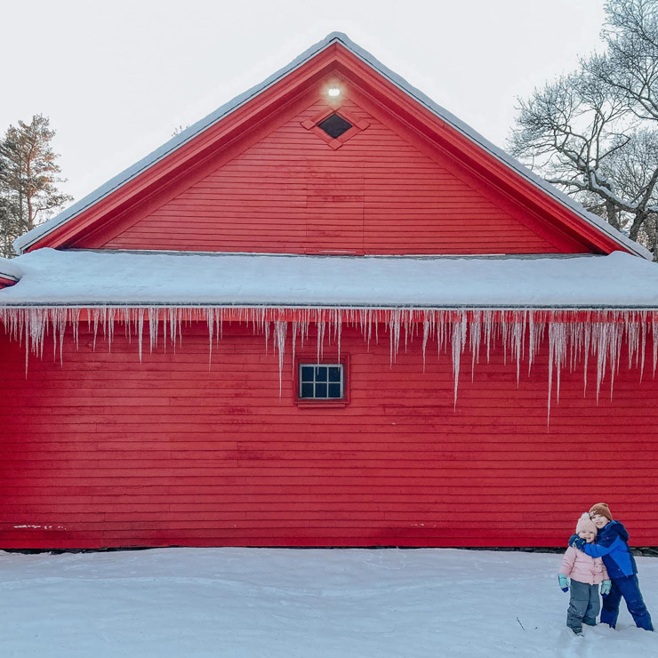 Winter scene of Claudia's children smiling in front of a red barn with icicles
